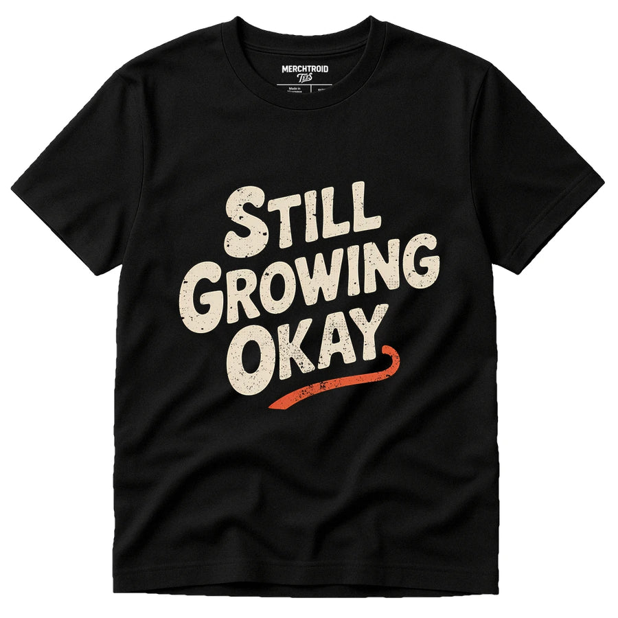 A black T-shirt with the phrase "STILL GROWING OKAY" printed in large, bold, off-white and red lettering on the front. The text has a slightly distressed, vintage look. The shirt is laid flat against a plain background.