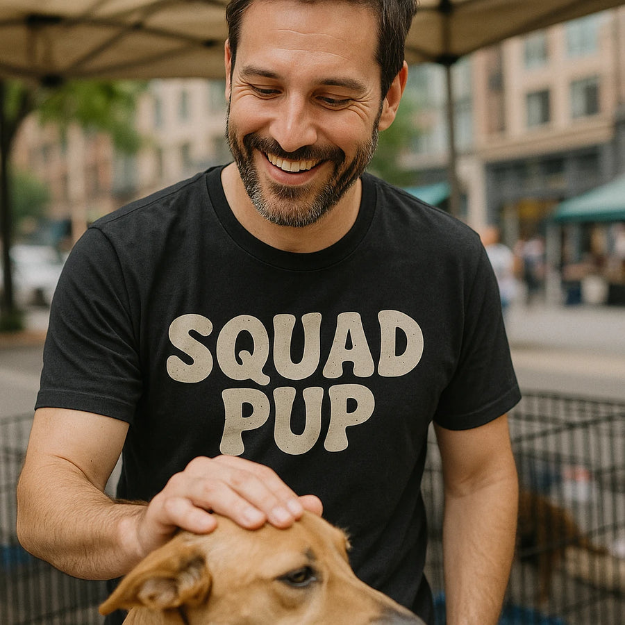 A smiling man with a beard is wearing a black t-shirt with the words "SQUAD PUP" printed on it. He is gently petting a light brown dog. They are in an outdoor setting with buildings and tents in the background.