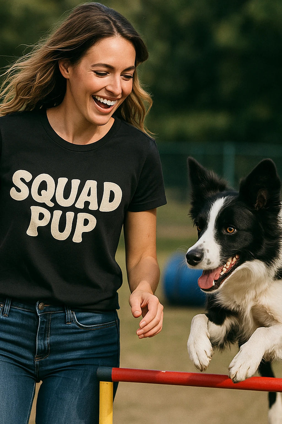 A person with long, wavy hair is smiling brightly while wearing a black t-shirt that says "SQUAD PUP" in bold letters. They are outdoors with a black and white dog enthusiastically jumping next to them, appearing to be engaging in an agility activity. The background is a blurred lawn and fenced area, suggesting a park or dog play area.