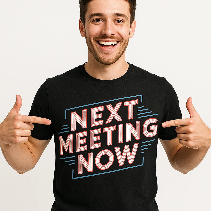 A person is smiling and pointing at their black t-shirt, which has the text "NEXT MEETING NOW" in bold, colorful letters.