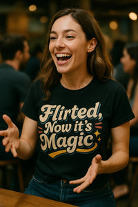 A person with long brown hair is smiling widely and gesturing with their hands. They are wearing a black t-shirt with colorful text. The background is blurred, with other people sitting around.