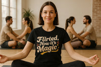 A person with short brown hair, wearing a black T-shirt with colorful text, is sitting cross-legged in a yoga pose, smiling at the camera. In the background, two couples sit facing each other, also cross-legged on yoga mats. The setting appears to be a bright, spacious room with a large window and a plant visible.