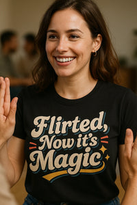 A person with long brown hair smiles while wearing a black t-shirt with colorful, whimsical text. The background shows blurred people sitting and talking.
