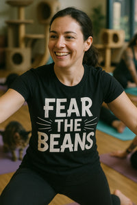 A person with long dark hair tied back is smiling during a yoga class. They are wearing a black t-shirt with the text 