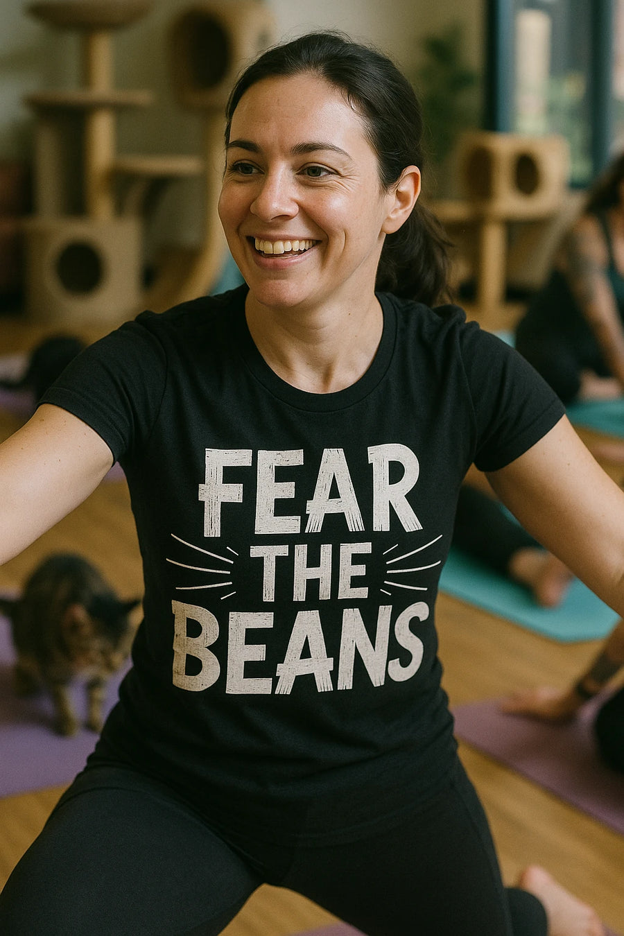 A person with long dark hair tied back is smiling during a yoga class. They are wearing a black t-shirt with the text "FEAR THE BEANS." In the background, there is a cat tree and a cat on a yoga mat. Other participants are visible behind them on yoga mats.