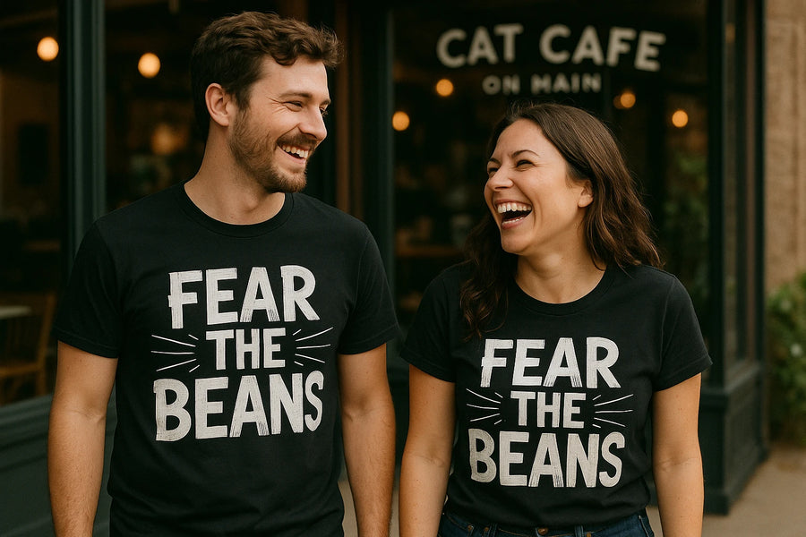 A man and a woman are standing outside a cafe, both wearing black t-shirts with "FEAR THE BEANS" printed on them. They are smiling and appear to be enjoying each other's company. The background shows a glass entrance with the words "CAT CAFE ON MAIN" visible.