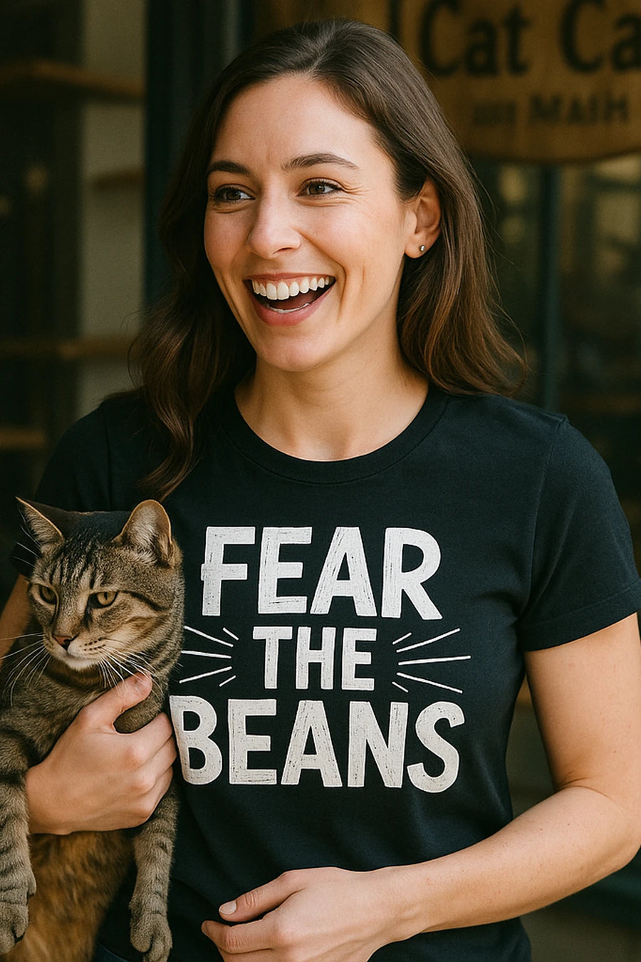 A person with long brown hair is smiling widely while holding a cat. They are wearing a black t-shirt that says "FEAR THE BEANS." The background shows a partially visible sign reading "Cat C...".